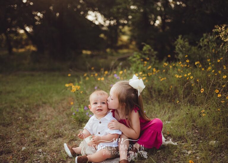 sister kissing baby brother outside near flowers