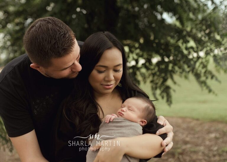 mom and dad holding newborn baby and looking down smiling for newborn photo session